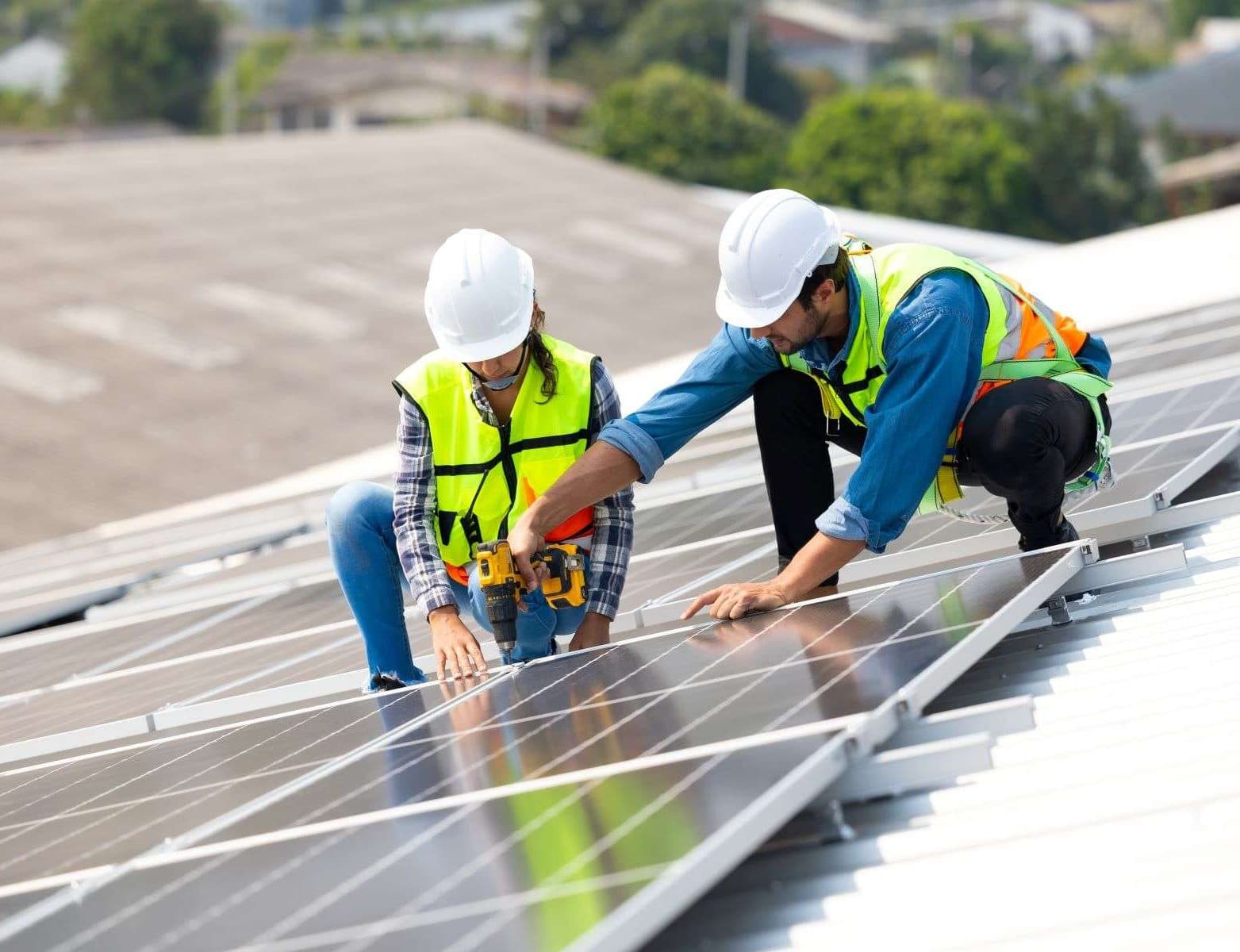 Two construction workers, a woman and a man, kneel down to work on a solar panel array.
